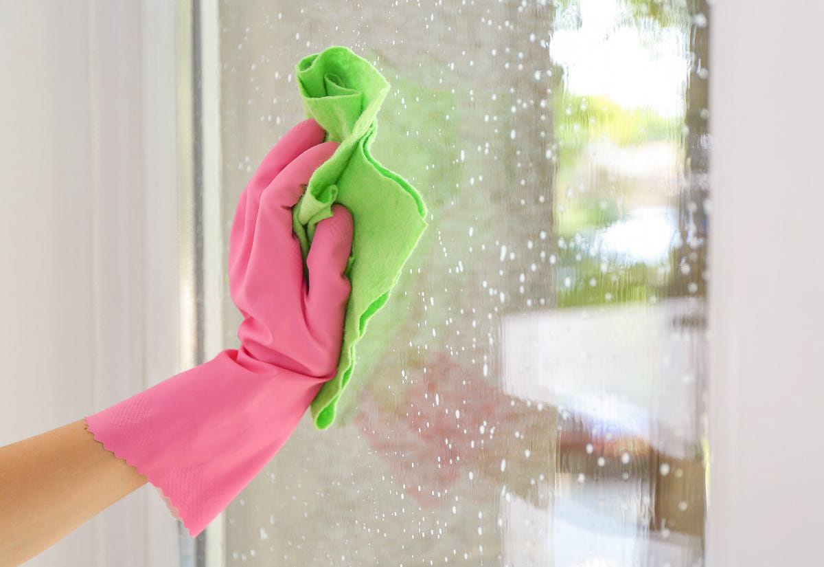 Hand wearing a pink rubber glove wiping a window with a green cloth while natural cleaning solution drips down the glass.