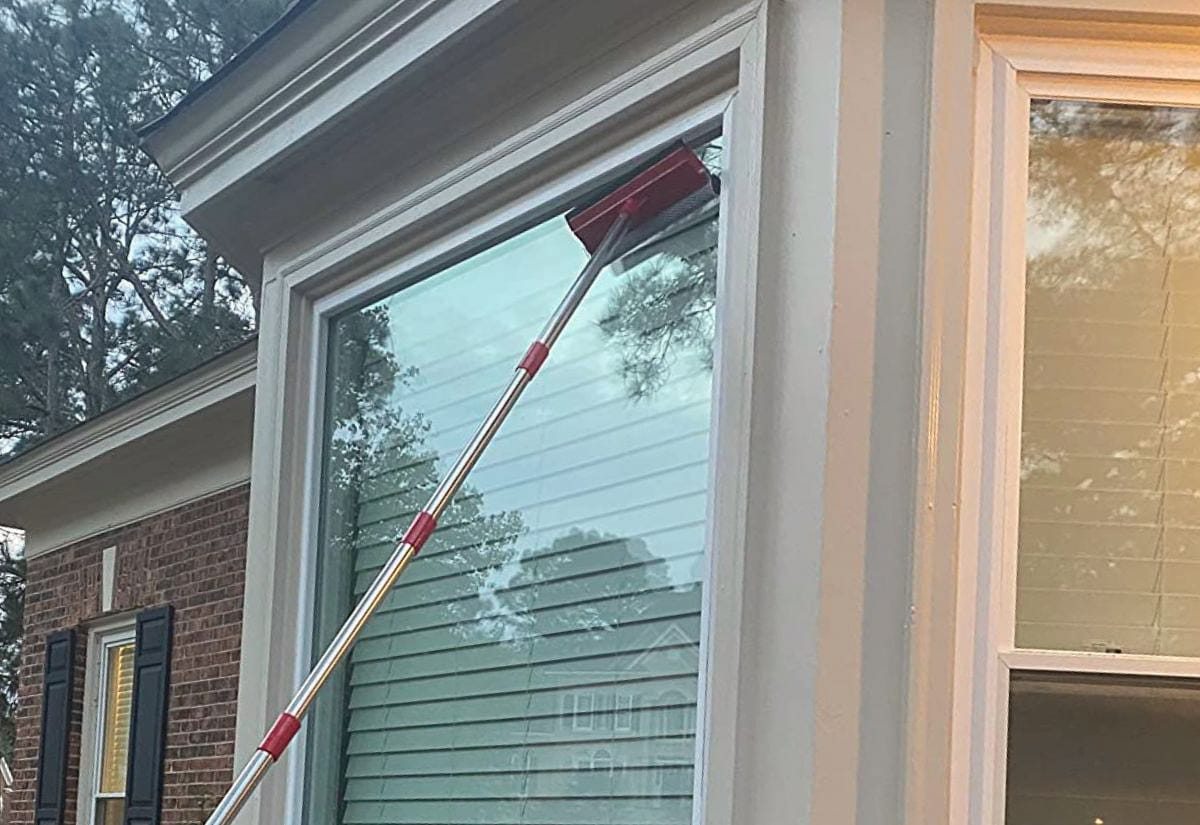 Exterior window being cleaned with a squeegee attached to an extension pole, showing streak-free glass on a home’s upper window.