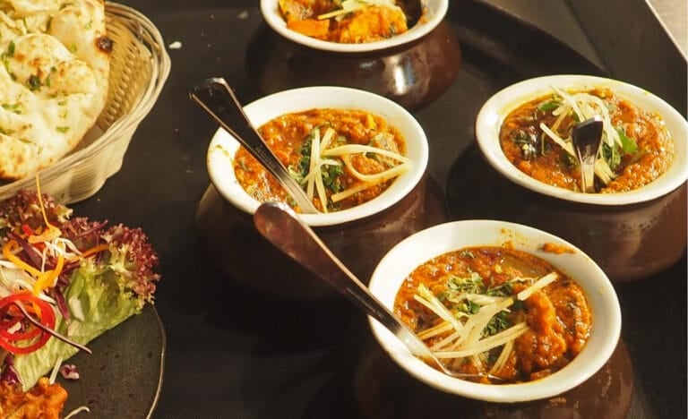 Four small ceramic bowls filled with hearty stew and topped with shredded vegetables, served with a fresh salad and naan bread on a dark tabletop.