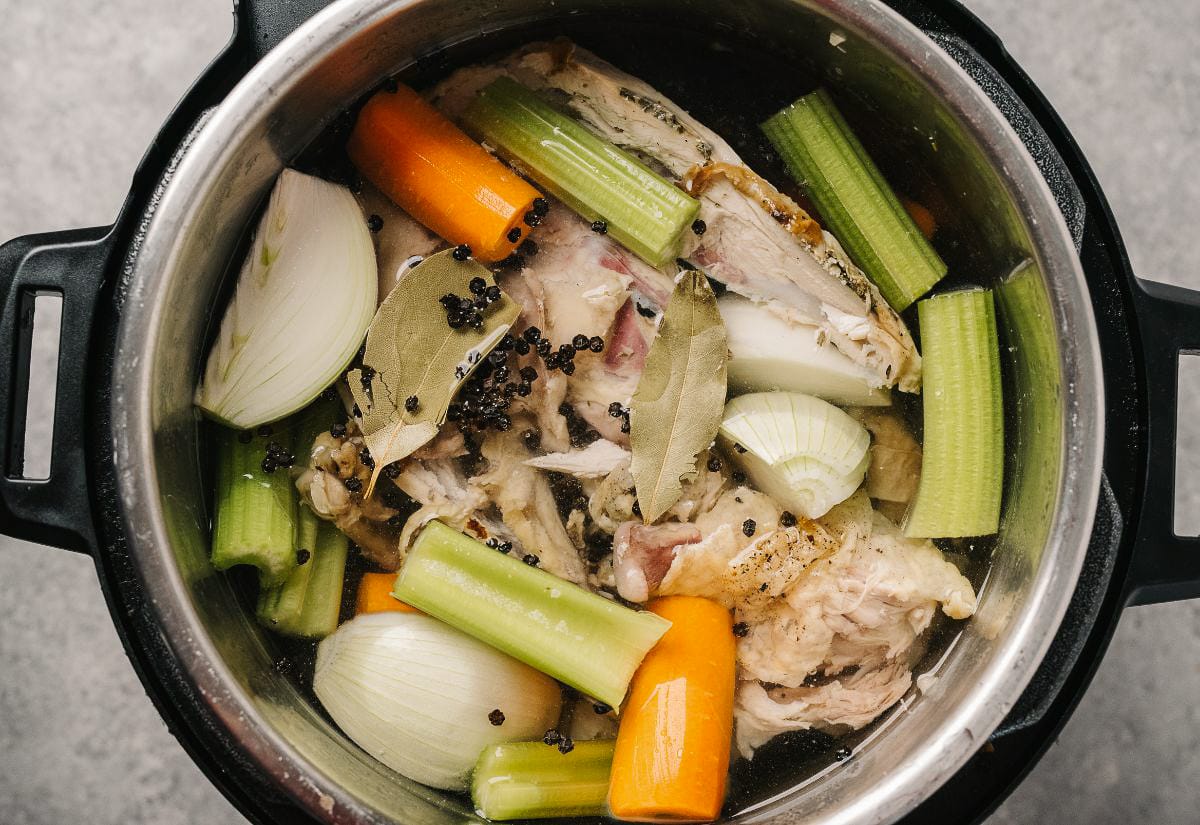 Close-up of an Instant Pot filled with homemade chicken broth ingredients including celery, onions, carrots, bay leaves, and peppercorns.