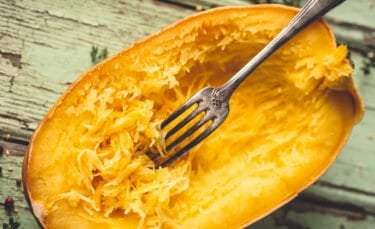Horizontal photo of a roasted spaghetti squash half with a fork raking through the strands, highlighting the spaghetti-like texture against a weathered mint-green background.