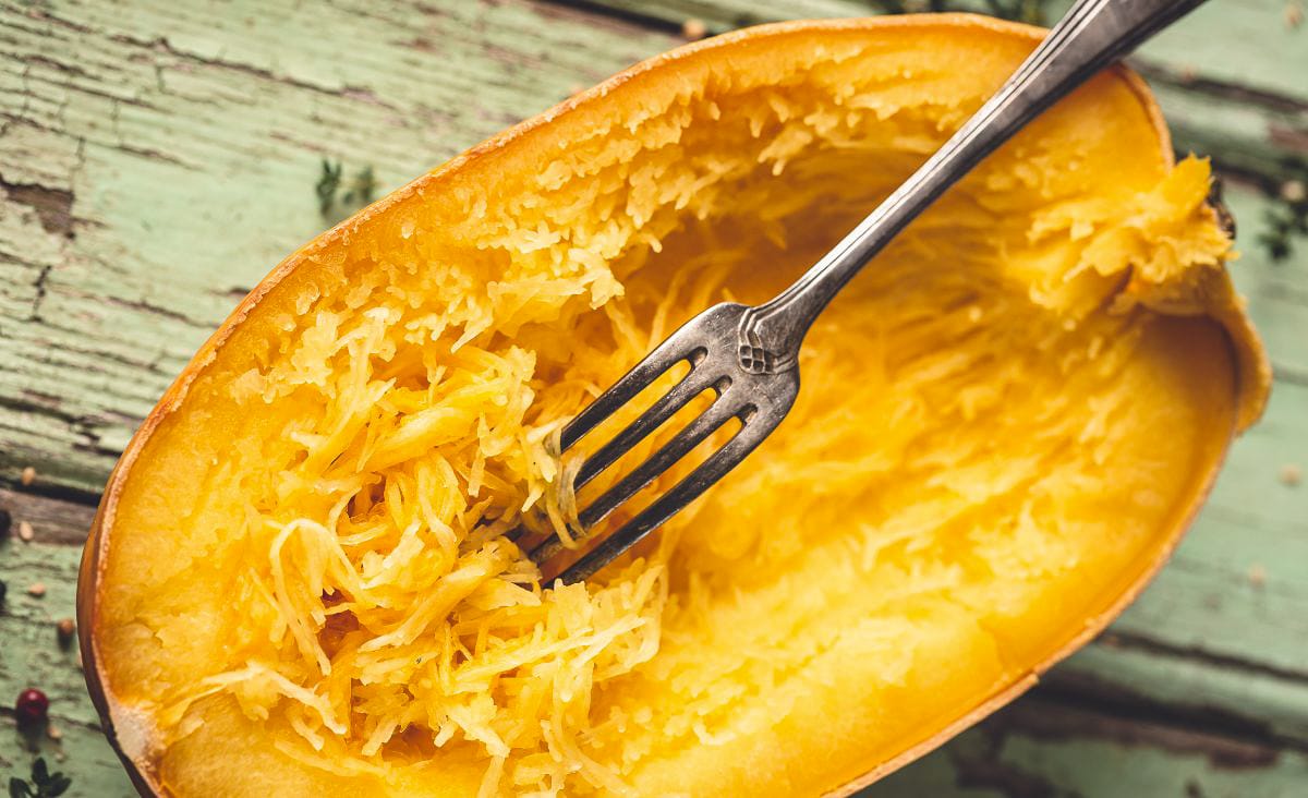 Horizontal photo of a roasted spaghetti squash half with a fork raking through the strands, highlighting the spaghetti-like texture against a weathered mint-green background.