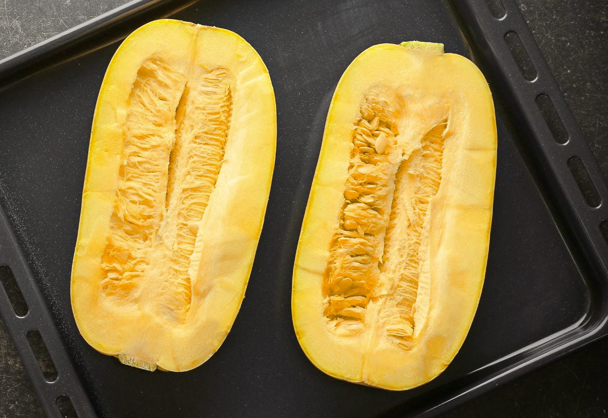 Two halved spaghetti squash placed cut-side up on a dark baking sheet before roasting, showing the fresh interior and visible seeds.