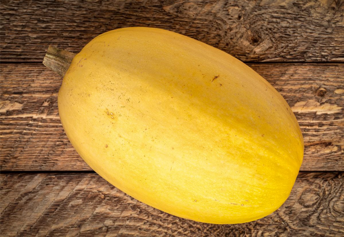 A whole uncooked spaghetti squash resting on a wooden table, showing its smooth yellow rind and natural imperfections.