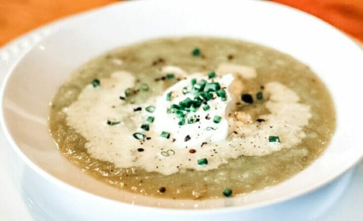 A closeup view of the potato, garlic, and leek soup in a shallow white bowl on an orange tablecloth. Soup is garnished with sour cream and chives.