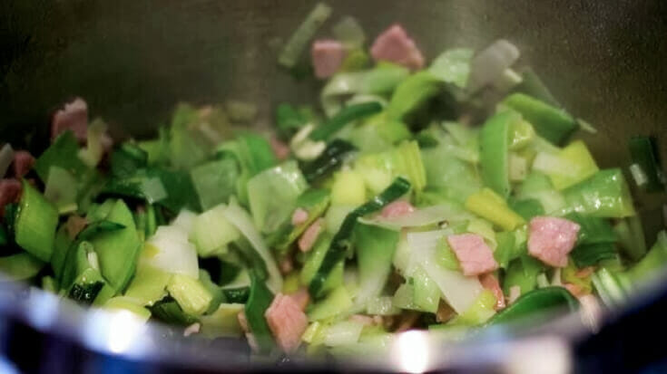 Chopped leeks, chopped bacon, and crushed garlic wilting in a stainless steel pot.