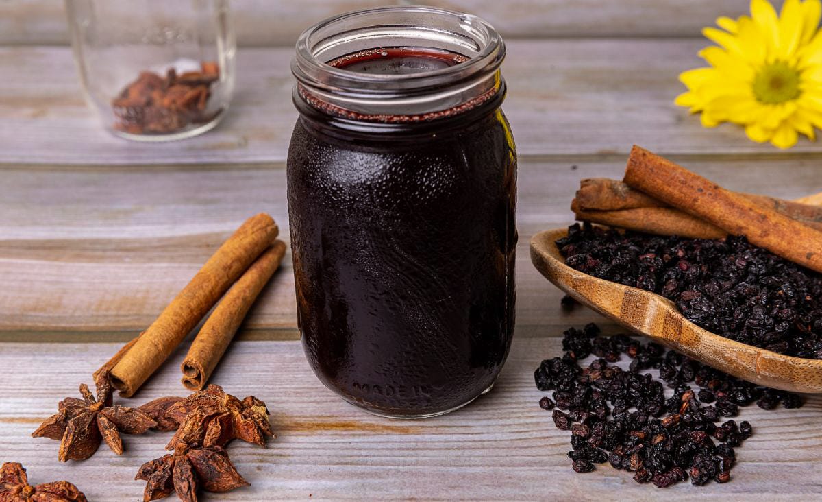 A close-up of a mason jar filled with dark elderberry syrup, set on a wooden table with cinnamon sticks and dried elderberries scattered nearby.