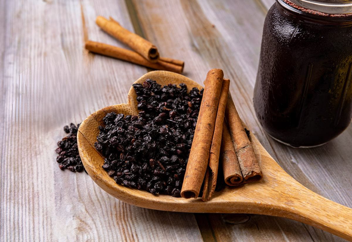 Dried elderberries fill a heart-shaped wooden spoon next to several cinnamon sticks, arranged on a weathered wooden surface for a cozy, handmade feel.