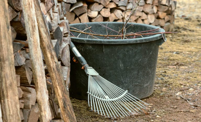A metal leaf rake resting against a large outdoor bin beside stacked firewood, showing a garden cleanup area ready for fall yard work.