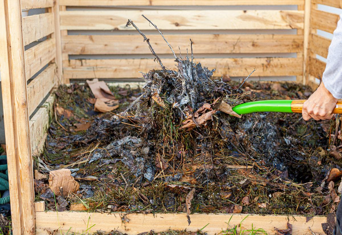 A hand using a green-handled garden fork to turn a compost pile filled with wet leaves, grass, and garden debris inside a wooden compost bin.