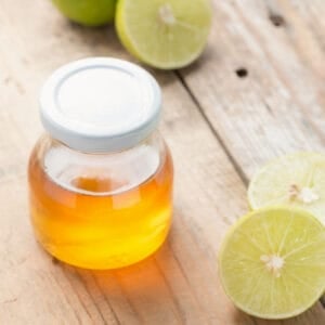 A glass jar filled with honey beside halved limes on a rustic wooden surface.