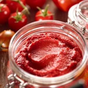 Glass jar filled with thick homemade tomato paste, deep red in color with a smooth texture, surrounded by fresh tomatoes in the background.