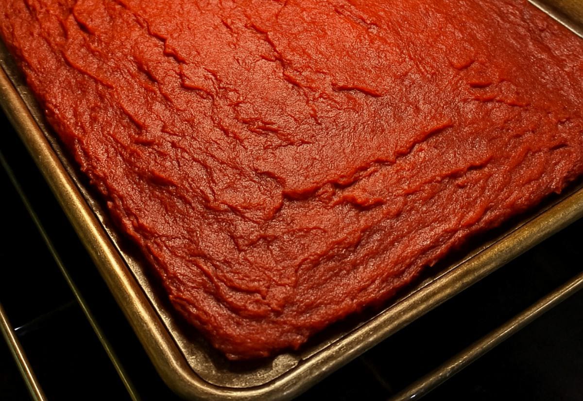 Brick-red homemade tomato paste baked down on a rimmed sheet pan in the oven, thick and glossy with visible cracks from reduction.