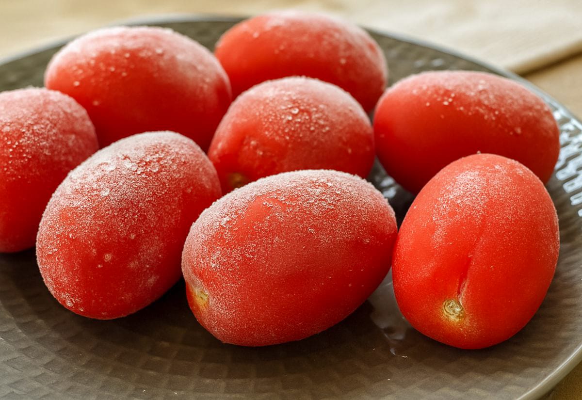 A plate of whole frozen Roma tomatoes with visible frost and ice crystals on the skins, just taken from the freezer before peeling.