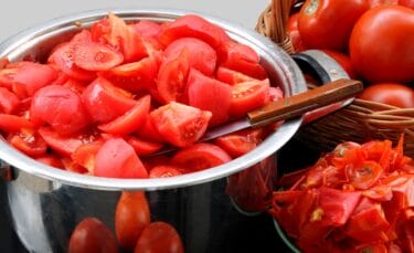 A steel mixing bowl filled with peeled tomatoes beside a plate of tomato peels.
