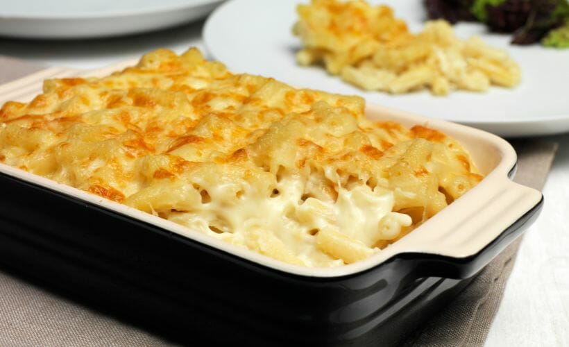 Baked macaroni and cheese in a black and white casserole dish with a plate of macaroni in the background.