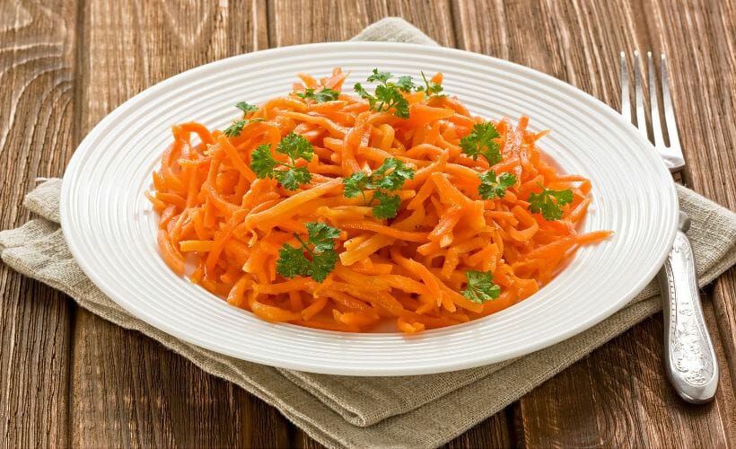 Fresh Russian carrot salad, topped with fresh parsley, in a white bowl on a wooden table.