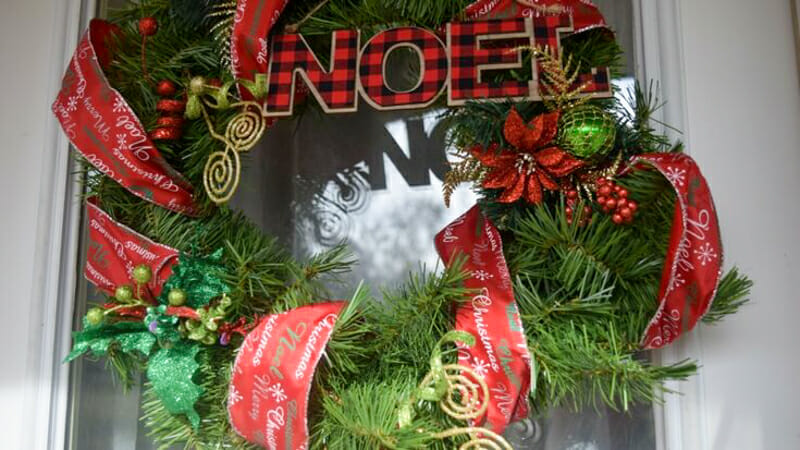 A wreath with christmas ribbon, ornaments, and a chipboard "noel" hanging from the front door.