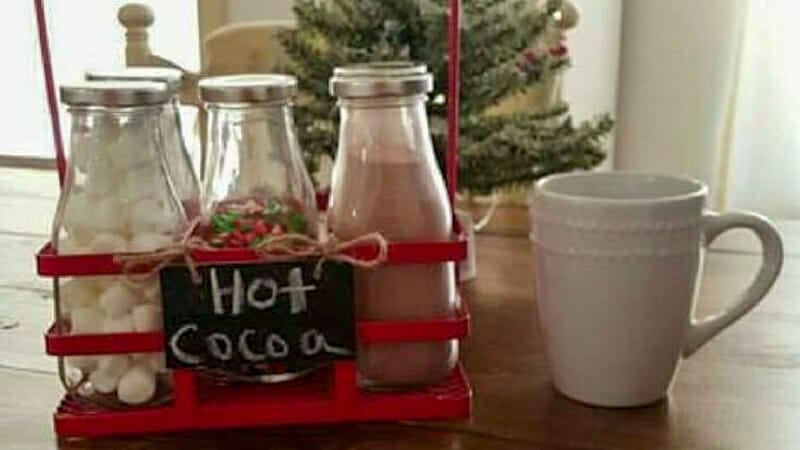 Cocoa fixings in vintage milk bottles in a red metal carrying crate sitting next to a white mug.
