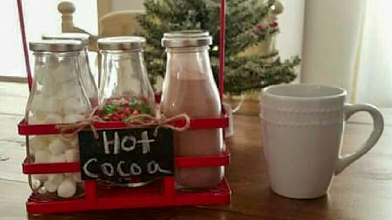 Cocoa fixings in vintage milk bottles in a red metal carrying crate sitting next to a white mug.