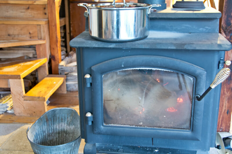 The woodstove with a pot simmering on top and the ash bucket beside it.