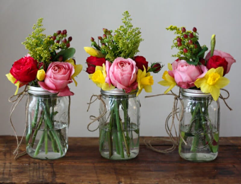 Roses, daffodils, carnations, and greenery arranged in three mason jars with twin tied around the tops.