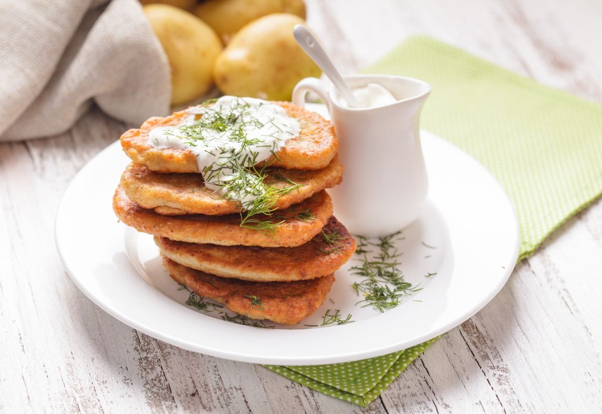 A stack of potato pancakes served with a pitcher of sour cream, fresh dill garnish, and whole potatoes in the background.