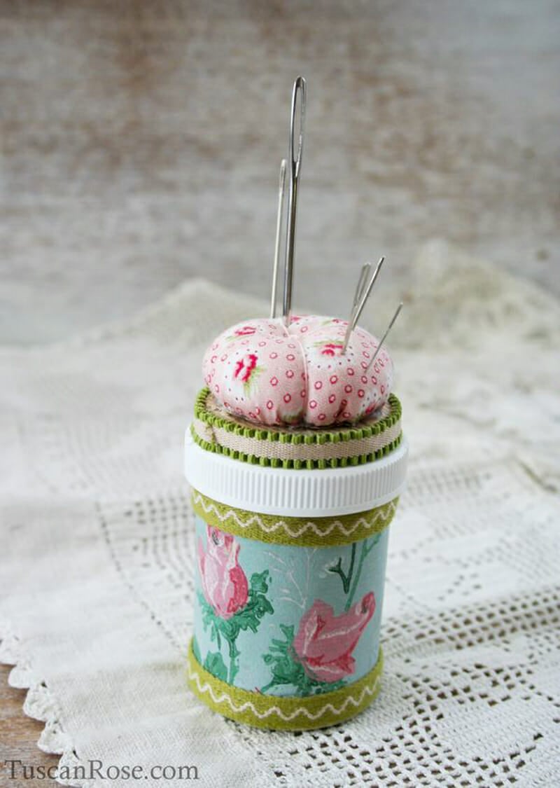 A pill bottle covered in rose fabric and ribbon with a pin cushion on top.