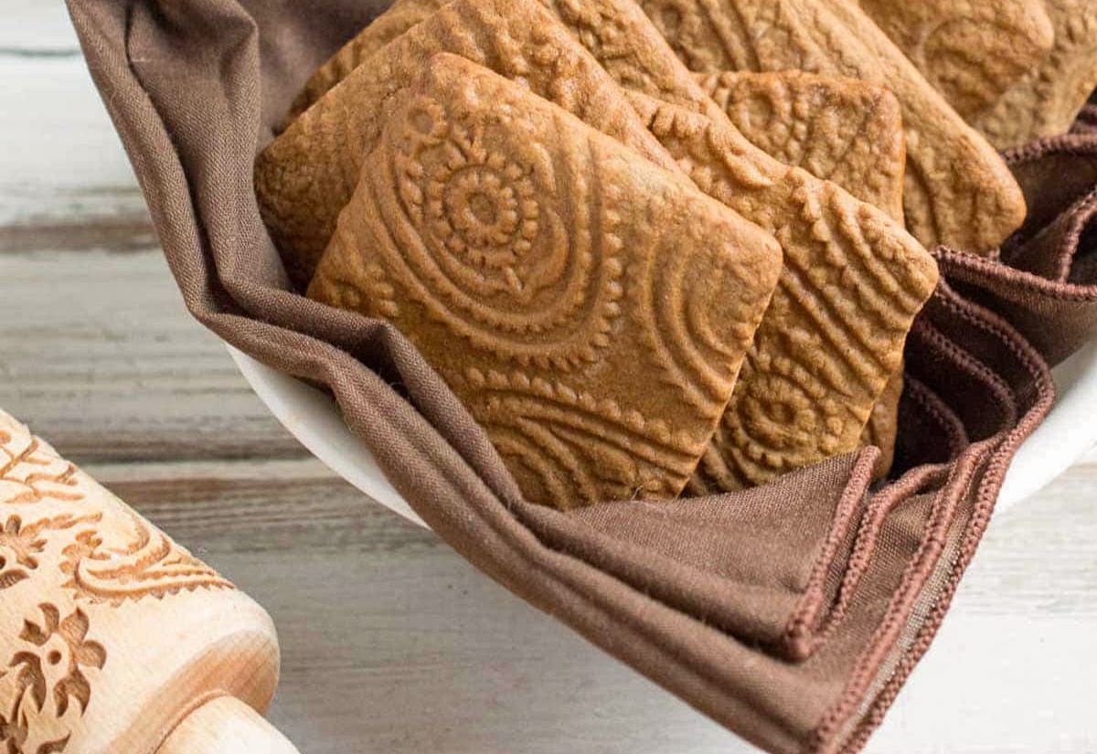 A shallow bowl lined with brown cloth holding square Speculaas cookies, each one stamped with ornate carved patterns that show off their detailed spice-cookie design.