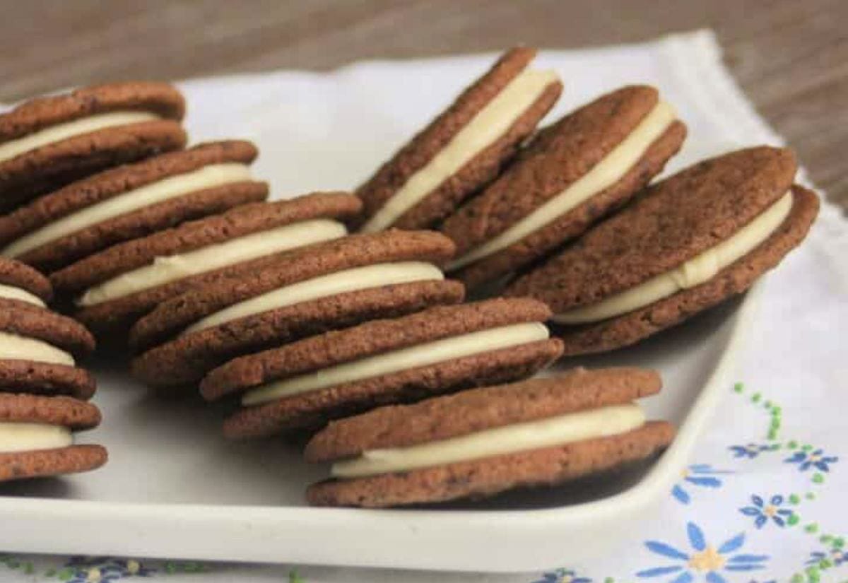 Thin chocolate sandwich cookies lined up on a white plate, each filled with a layer of pale vanilla buttercream, arranged neatly on a patterned cloth.