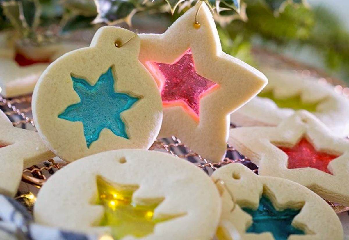 Cut-out sugar cookies in ornament and star shapes, each with a colorful “stained-glass” candy center glowing in shades of blue, yellow, and pink.