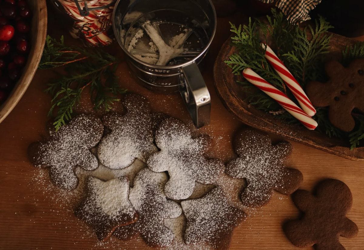 Soft gingerbread cookies in gingerbread-person and star shapes dusted with powdered sugar, surrounded by holiday greenery, candy canes, and a metal sifter on a wooden table.