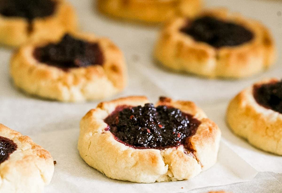 Rustic thumbprint cookies with golden, craggy edges and a dark mulberry jam center, baked on parchment paper.