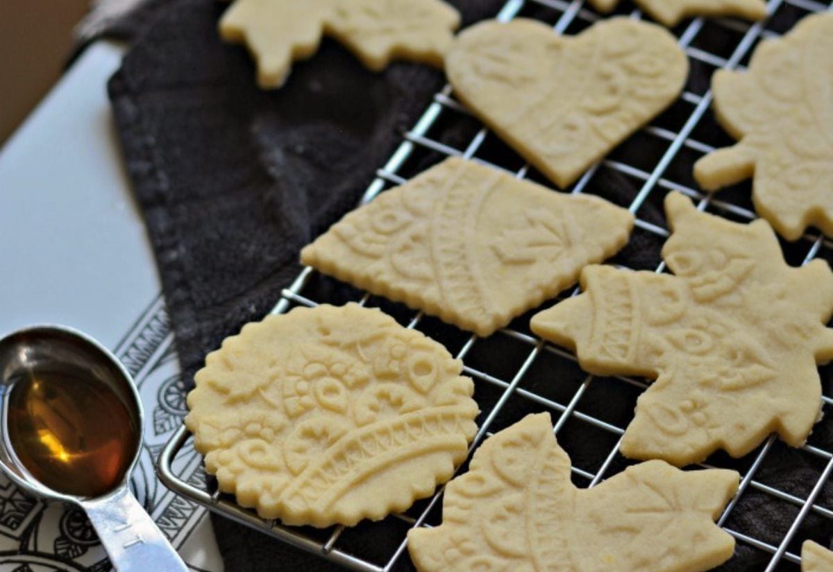 Embossed sugar cookies cooling on a metal rack, each cut into shapes like hearts, bells, and trees, with delicate stamped designs visible across the pale dough.