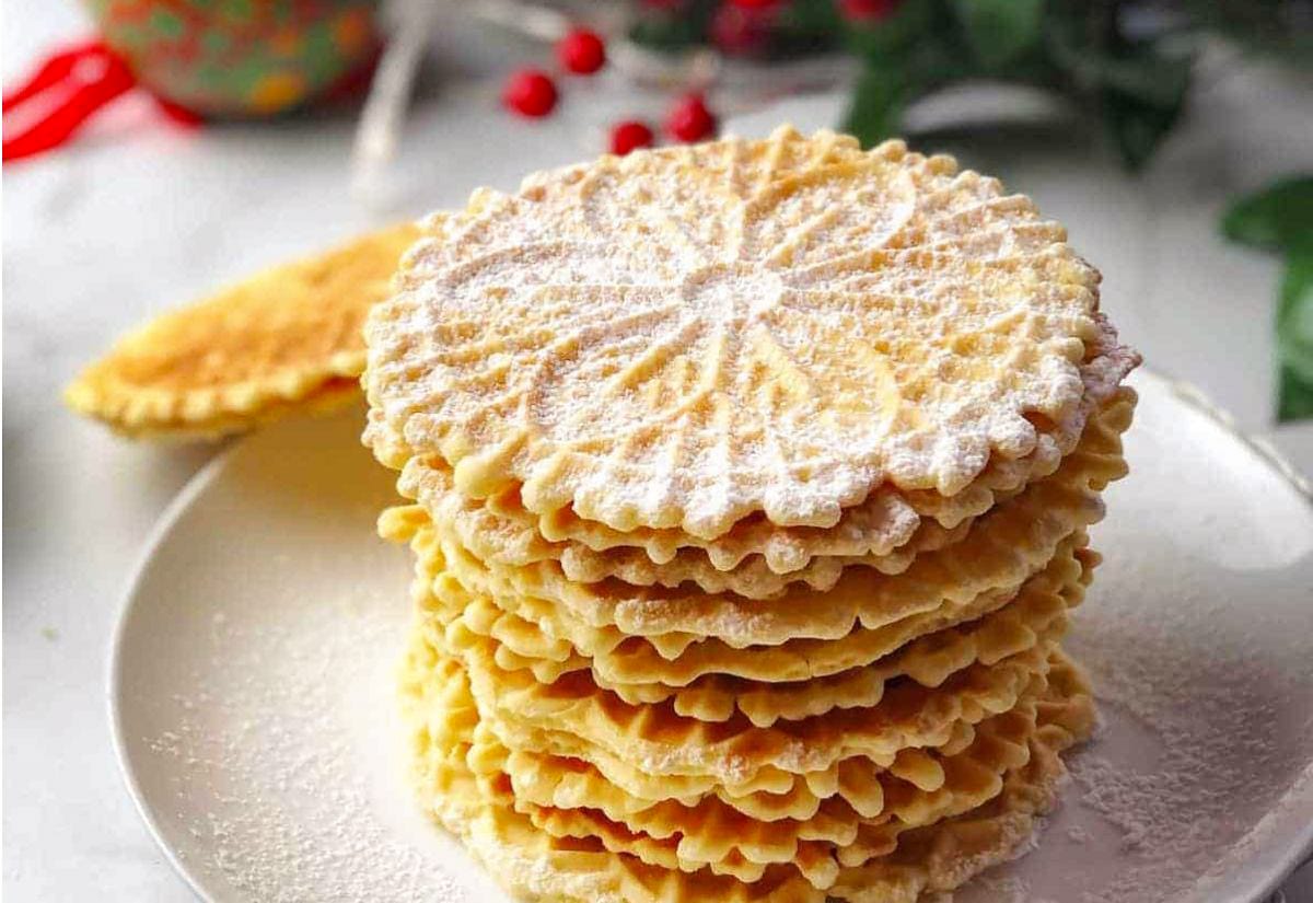 A tall stack of golden pizzelle cookies dusted with powdered sugar, showing off their thin, lace-like pattern on a white plate with holiday greenery in the background.