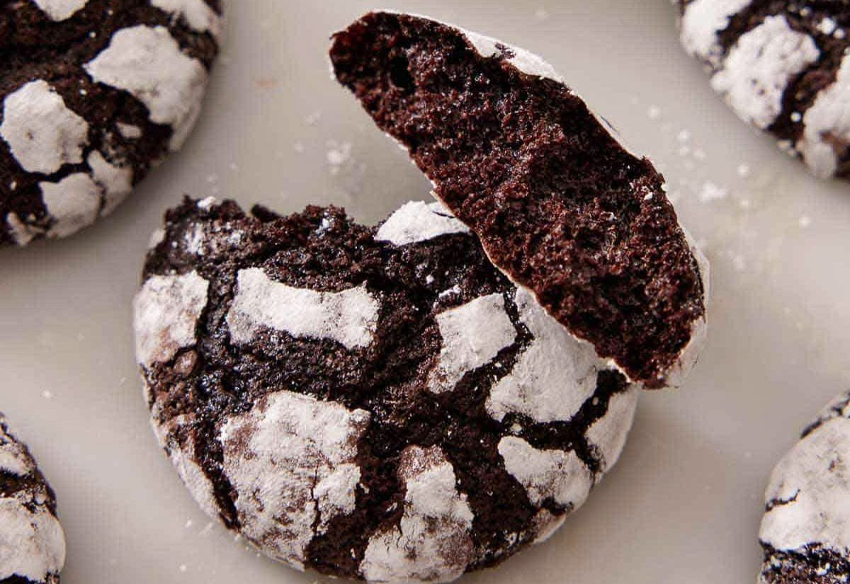Close-up of chocolate crinkle cookies on a baking sheet, one broken open to show the soft fudgy center beneath the cracked powdered sugar coating.
