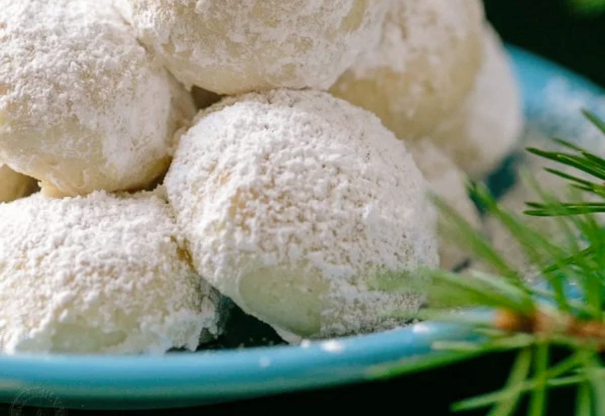 Powdered sugar–coated almond snowball cookies piled on a turquoise plate, with a sprig of evergreen peeking into the frame for a holiday touch.