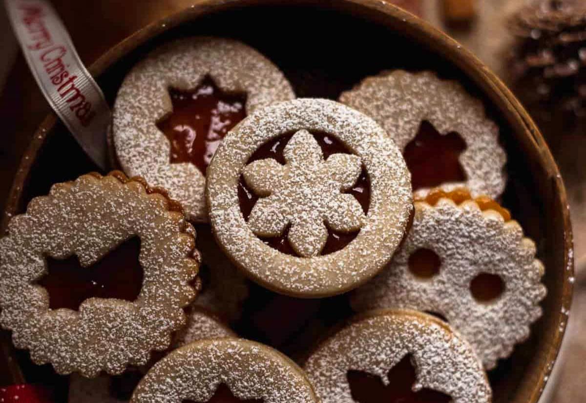A tin filled with classic Linzer cookies dusted with powdered sugar, each with a cut-out shape on top revealing the jam layer underneath.