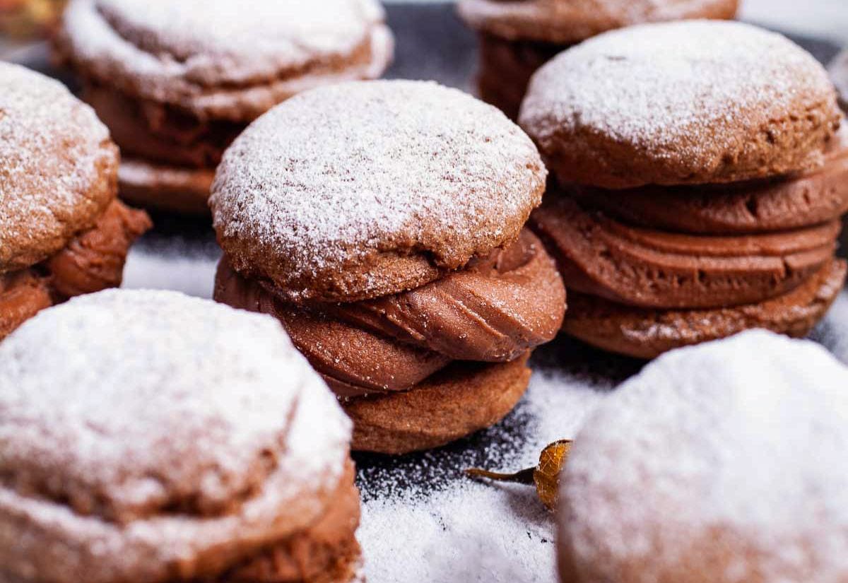 Soft chocolate sandwich cookies filled with thick chocolate frosting and dusted with powdered sugar, arranged closely together on a dark tray.