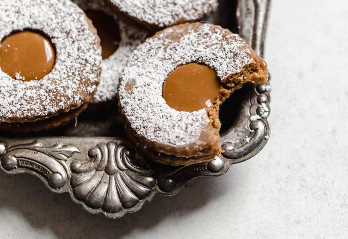 Round gingerbread-style Linzer cookies dusted with powdered sugar, each with a caramel-filled center; one cookie shows a bite taken out, displayed on a vintage silver tray.