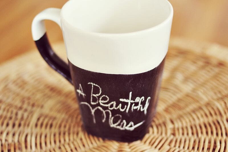 A white coffee cup with black chalkboard paint and "a beautiful mess" written on it.