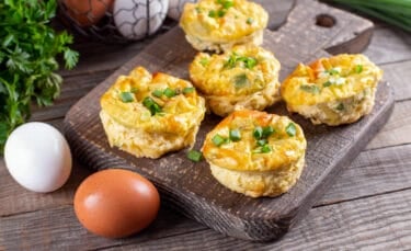 A rustic wooden board holding freshly baked mini quiches topped with chopped scallions, with brown and white eggs and fresh herbs in the background.