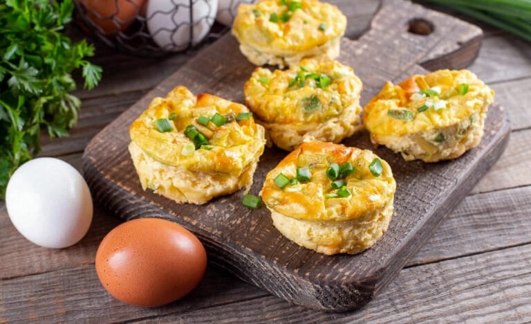 A rustic wooden board holding freshly baked mini quiches topped with chopped scallions, with brown and white eggs and fresh herbs in the background.