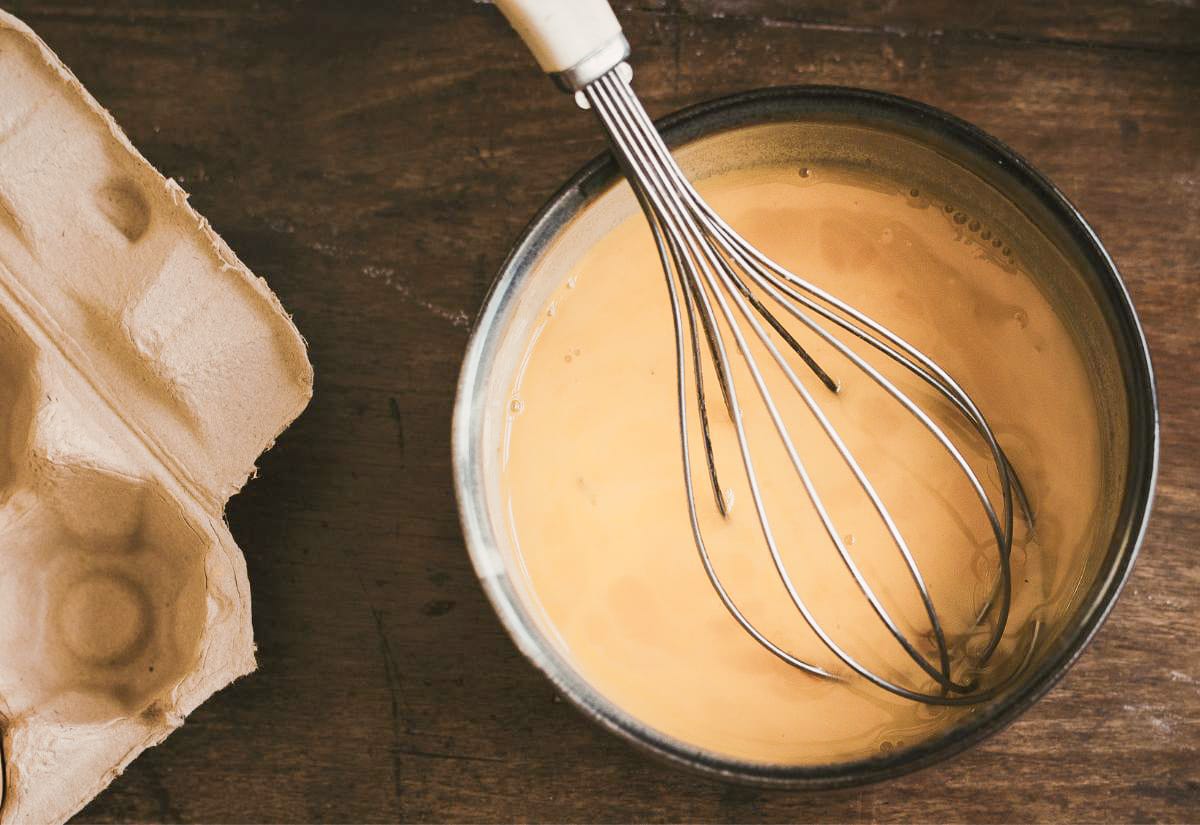 A bowl of whisked eggs mixed with milk and seasoning, with a metal whisk resting inside and an empty egg carton beside it.