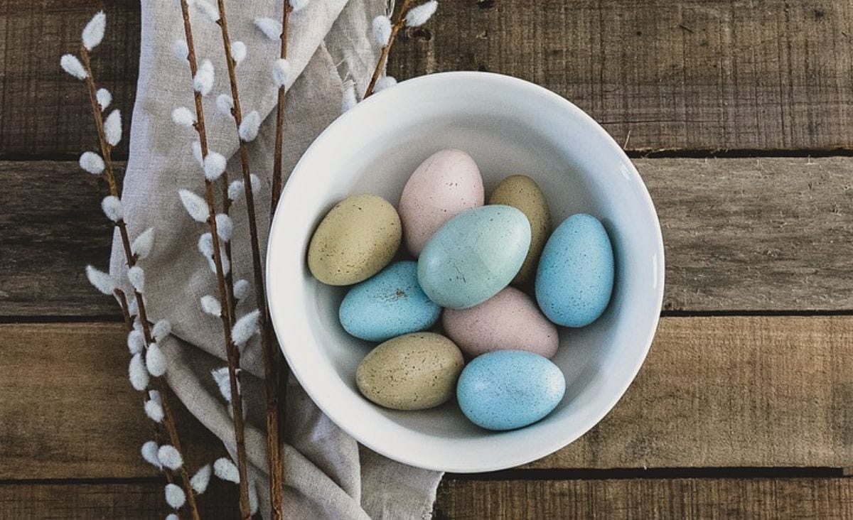 White bowl filled with naturally dyed Easter eggs in muted blue, pale pink, and olive green shades sitting on a rustic wooden table, with soft linen fabric and pussy willow branches arranged beside the bowl.