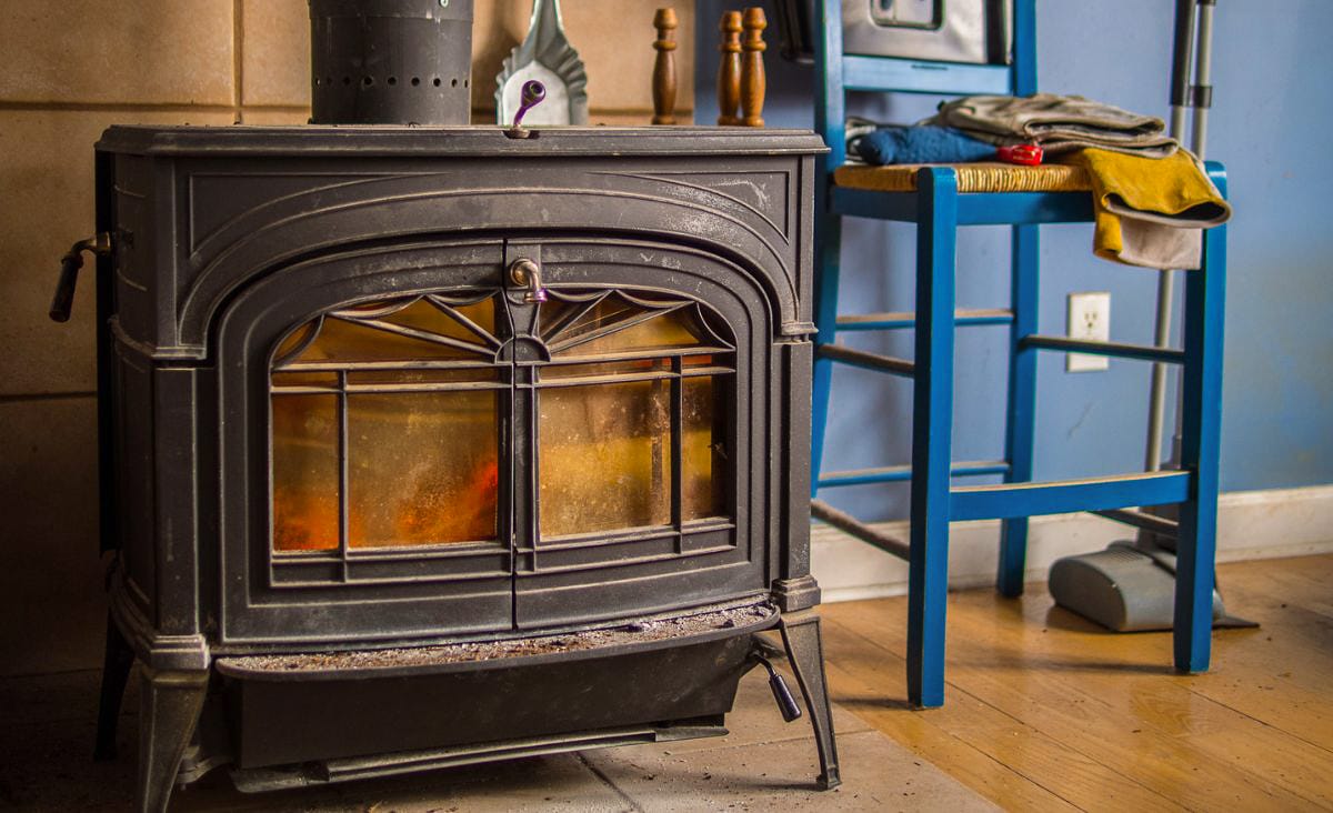 Close-up of a black wood stove with glowing embers behind the glass doors, set beside a blue chair and folded winter clothes in a rustic Maine home.