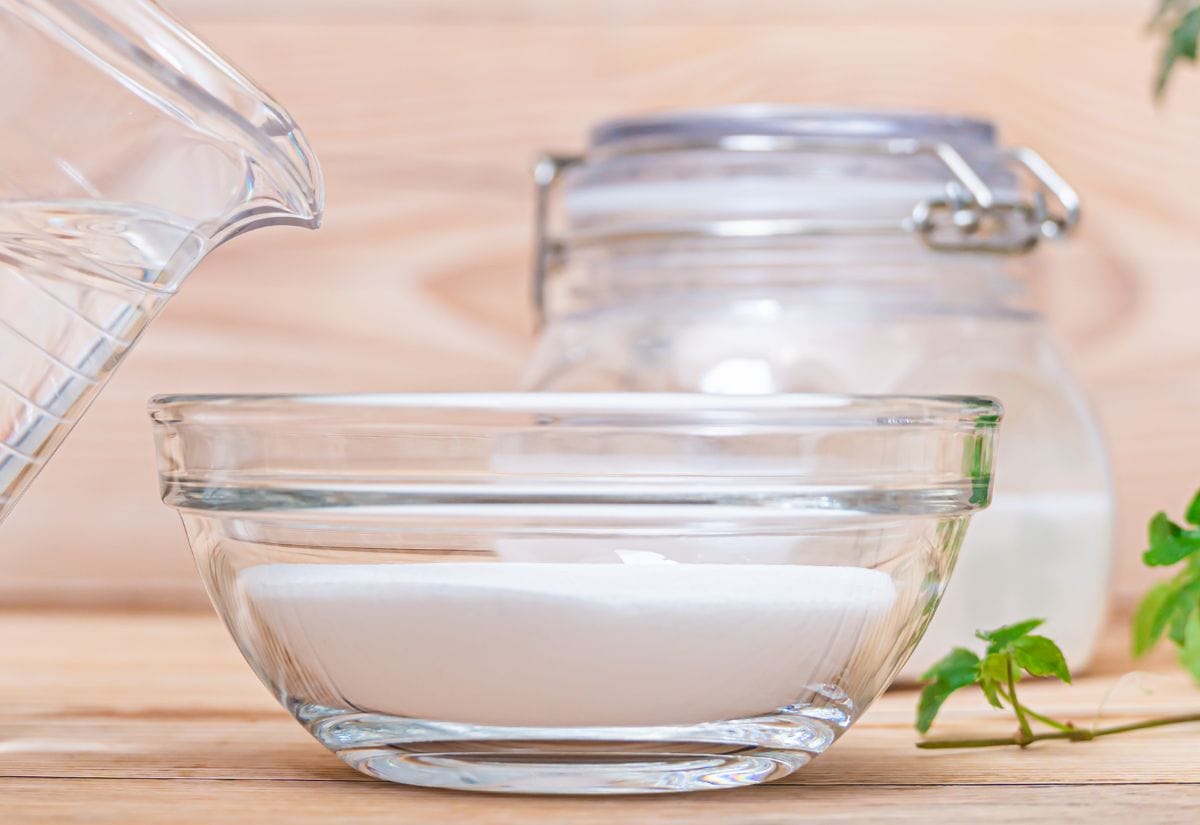 A glass bowl filled with baking soda and a jar of vinegar in the background, ready for cleaning use.