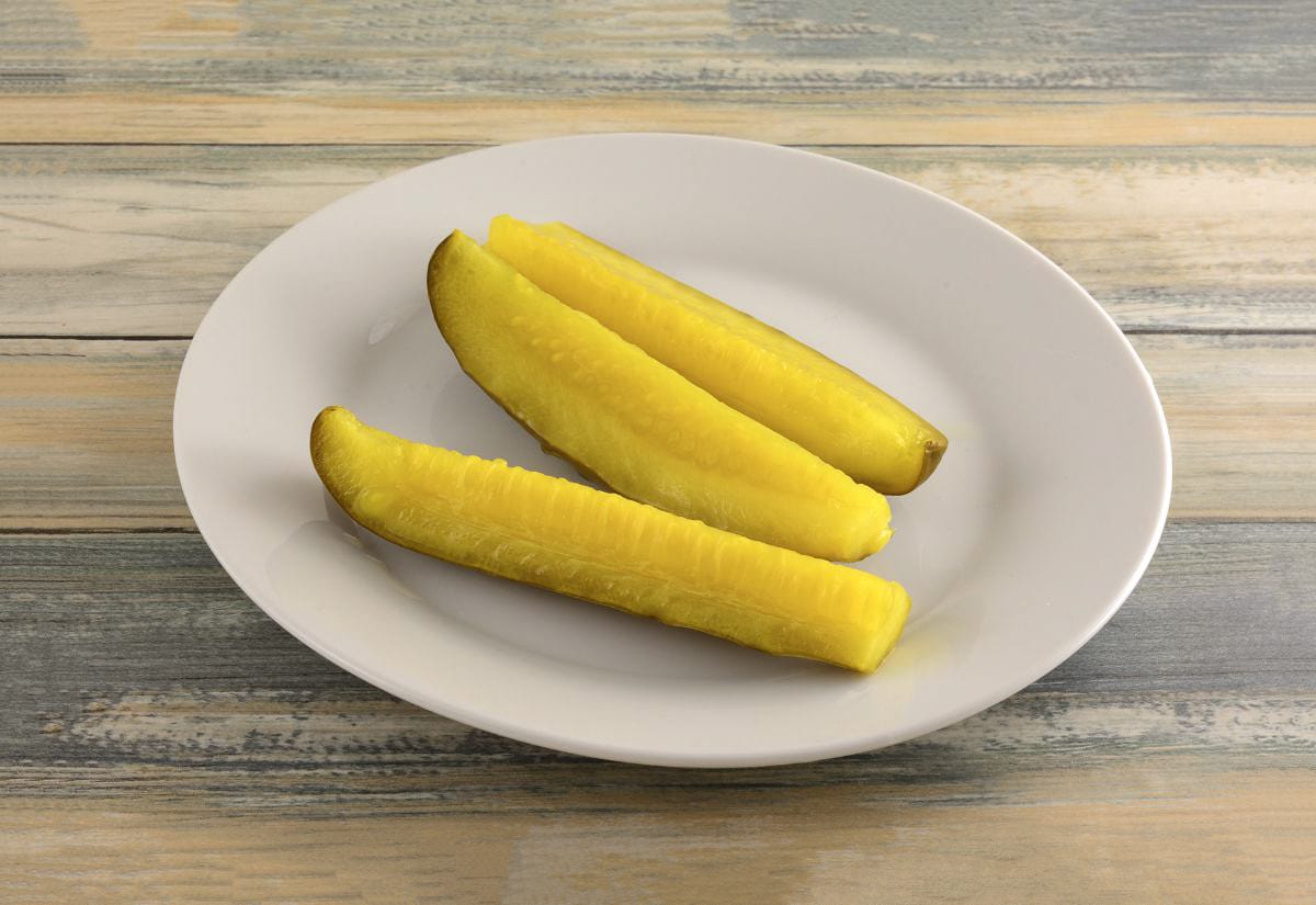 Three bright yellow-green dill pickle spears arranged neatly on a white plate. Their texture and sheen highlight their crispness and tangy flavor