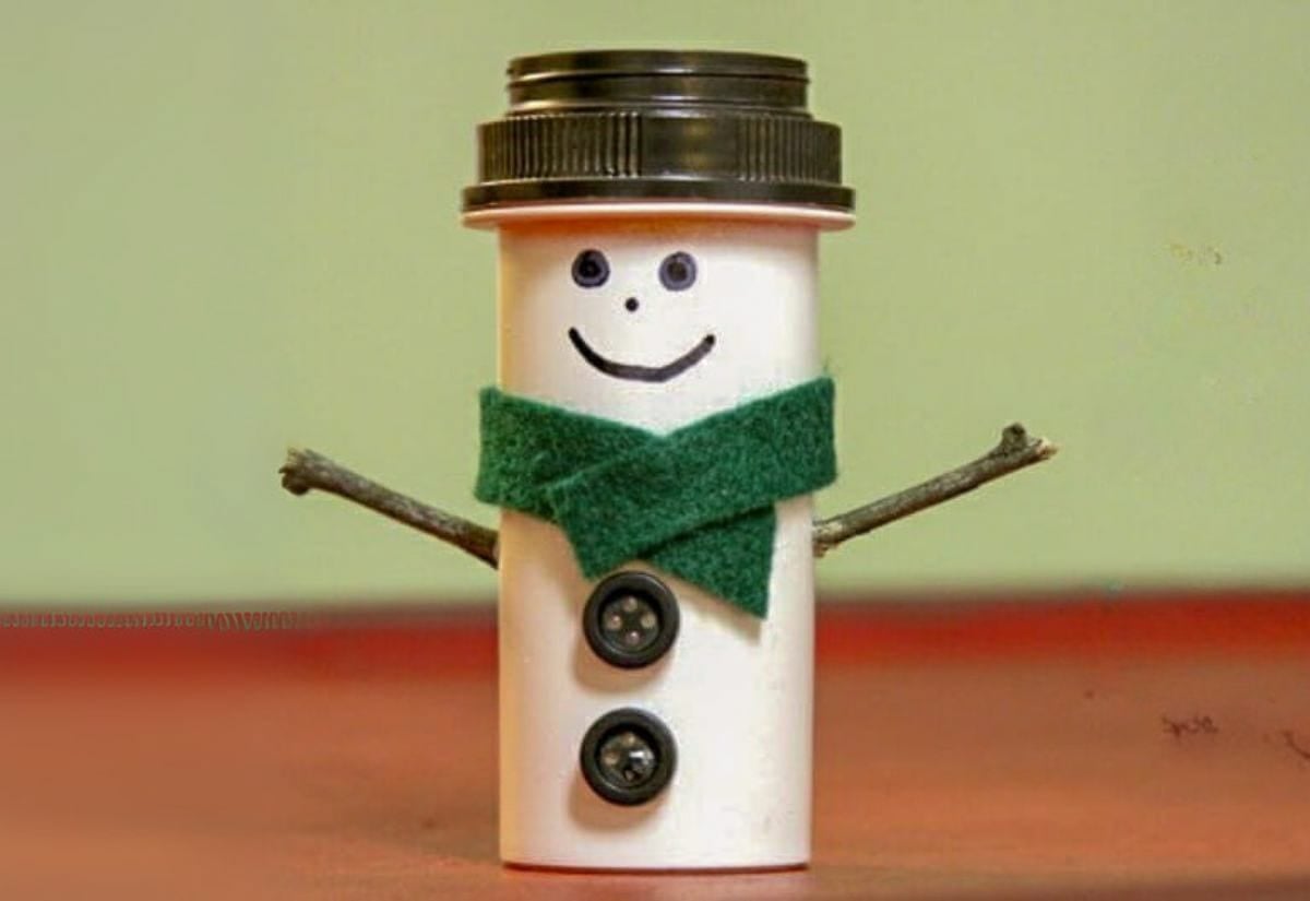 White pill bottle decorated as a snowman with a black hat, green felt scarf, stick arms, and button details.