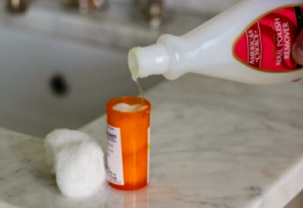 Bottle of nail polish remover being poured into a pill bottle stuffed with cotton balls, next to a cotton ball on a bathroom counter.